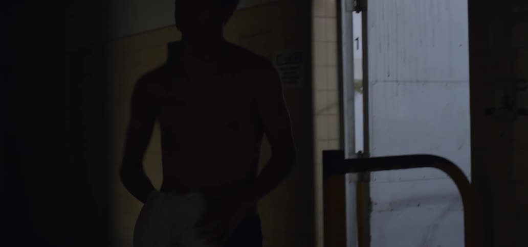 A young man pulls his shirt off as he enters and walks down a dark hallway