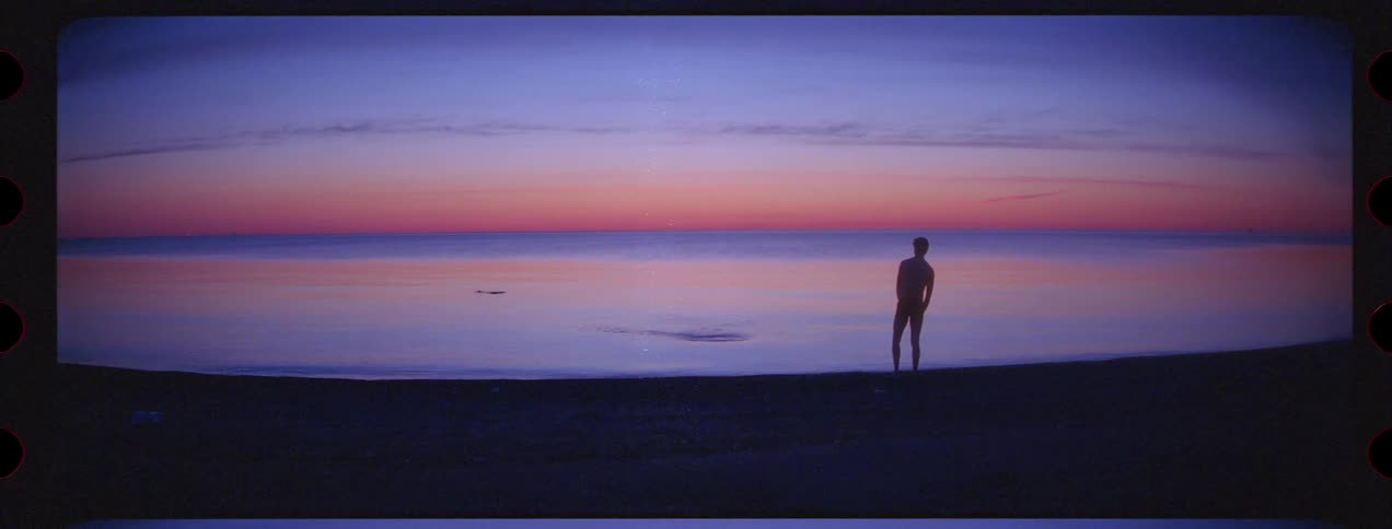 Silhouette of a man standing on the shoreline of a beach throwing pebbles at the sea at sunset