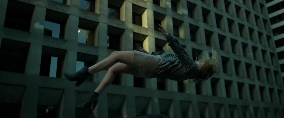 A woman in a jacket and boots falls to the ground in front of a concrete building