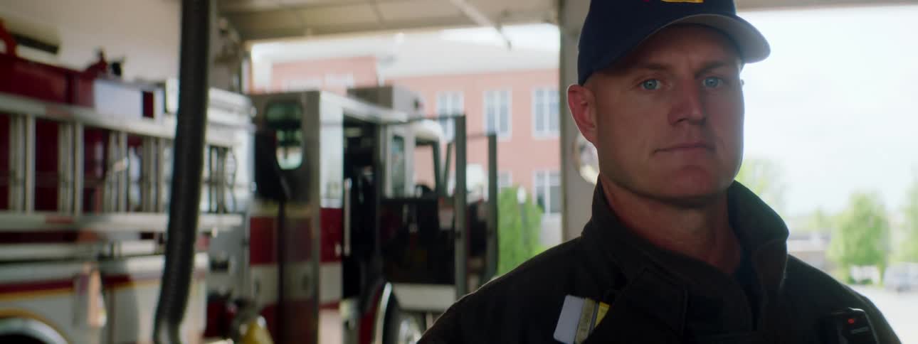 A man with a hat and jacket and a walkie talkie standing in a fire station with a fire truck behind him