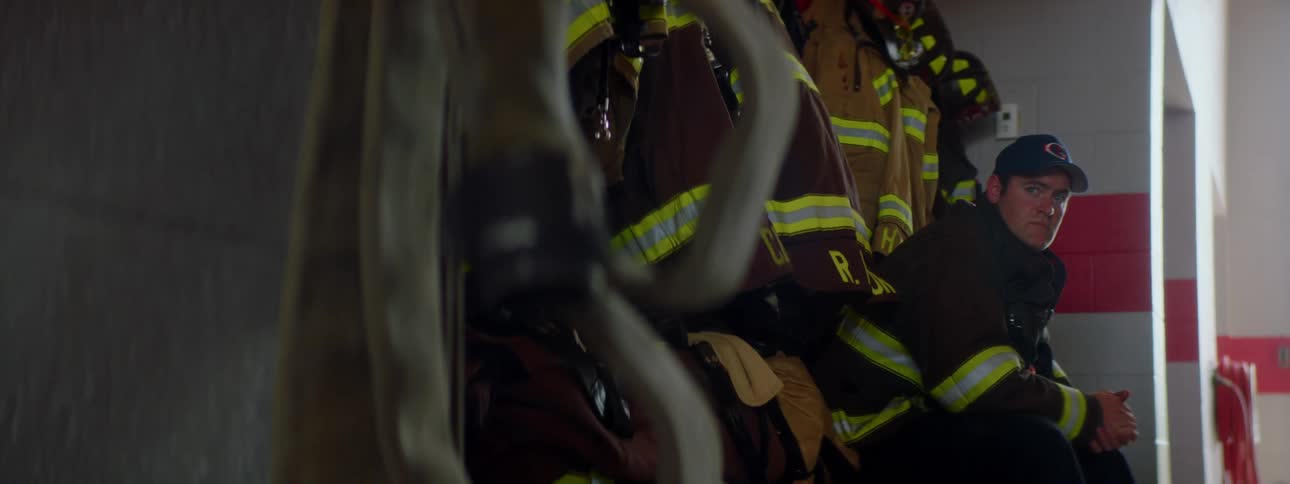 A man with a hat and a coat sitting down in a fire station with helmets and coats behind him