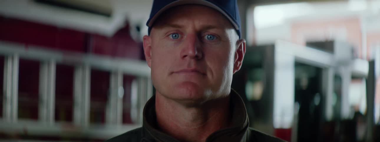 Firefighter with a blue cap and and coat standing next to a fire truck raises his head and looks at the camera in a fire station