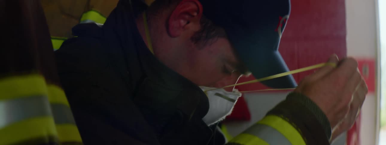 Firefighter with a cap and coat sits on a bench and wears a white mask in the fire station