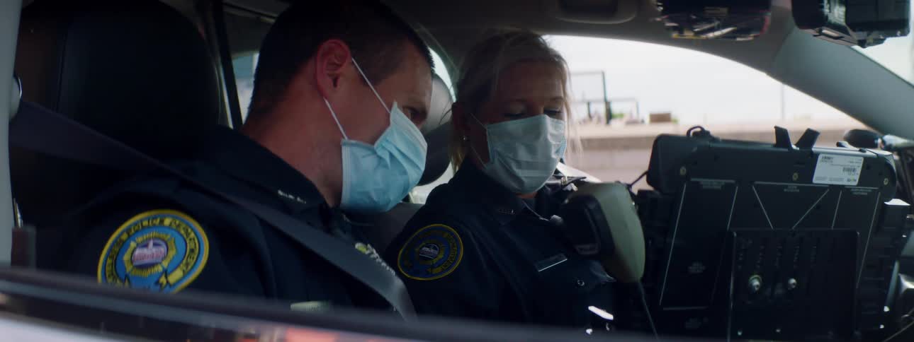 A male and female police officer in medical masks sit in a police car and look at the car computer