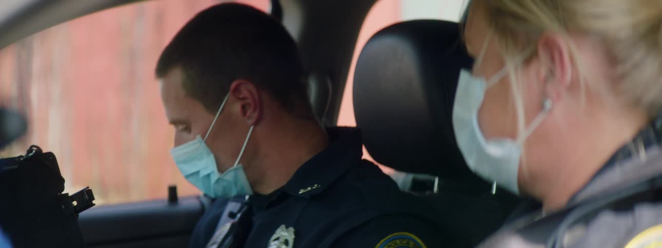 Two police officers sit inside of an idle police car as the male officer looks around the interior of the car