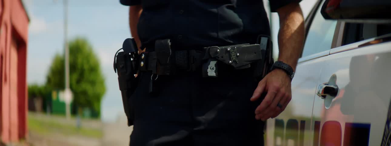 A police officer walking towards a police car and taking out gloves with trees utility poles and the sky in the background