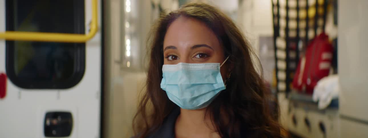 Woman with brown hair and a light blue mask faces the camera with eye contact