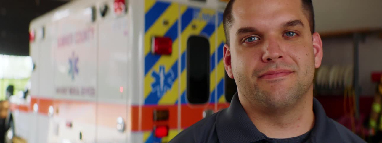 Close up of a mans face with blue eyes standing in front of an ambulance