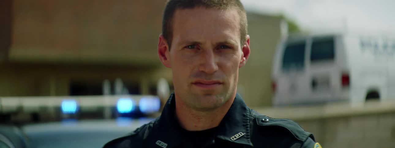 A police officer standing and staring with a police car a van and a building in the background