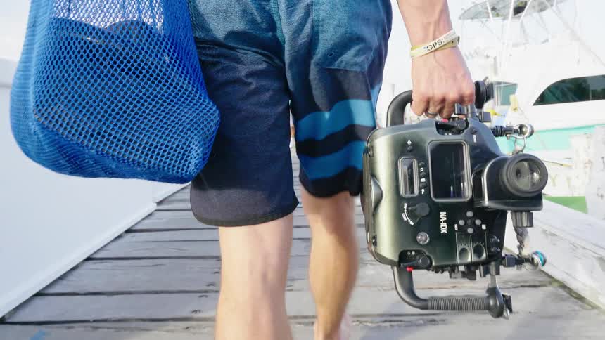 Man carries a mesh bag and a camera as he walks on a pier near the boats