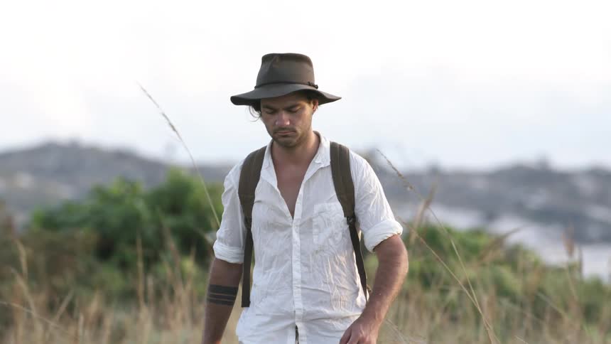 Man walks in a field next to a river while wearing a cowboy hat