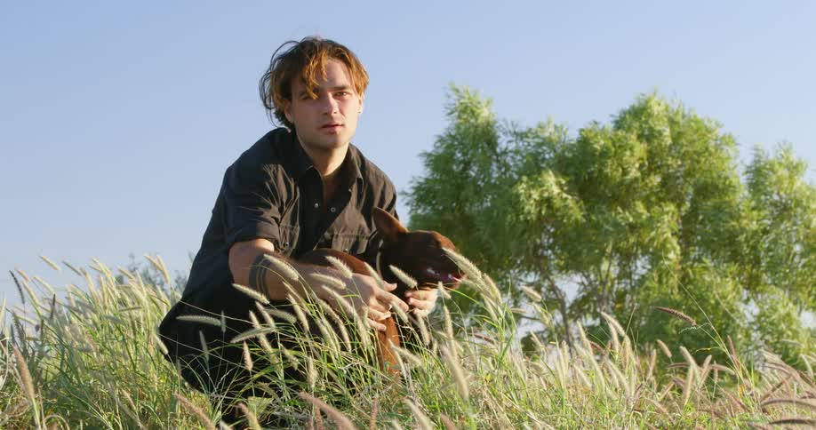A young man pets his small dog as he crouches in a grassy field outdoors