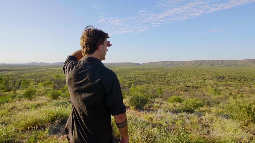 Man with a black shirt stands and looks around in an open field on a sunny day