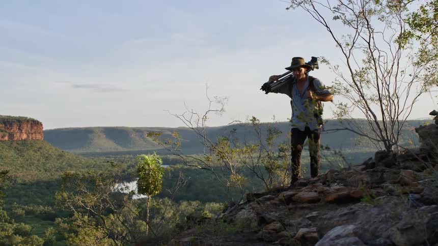 Photographer carries tripod and camera along top of slope with mesa and a river in the background