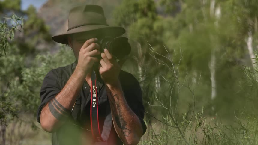 Photographer takes aim with camera standing in field near trees under a tall mountain