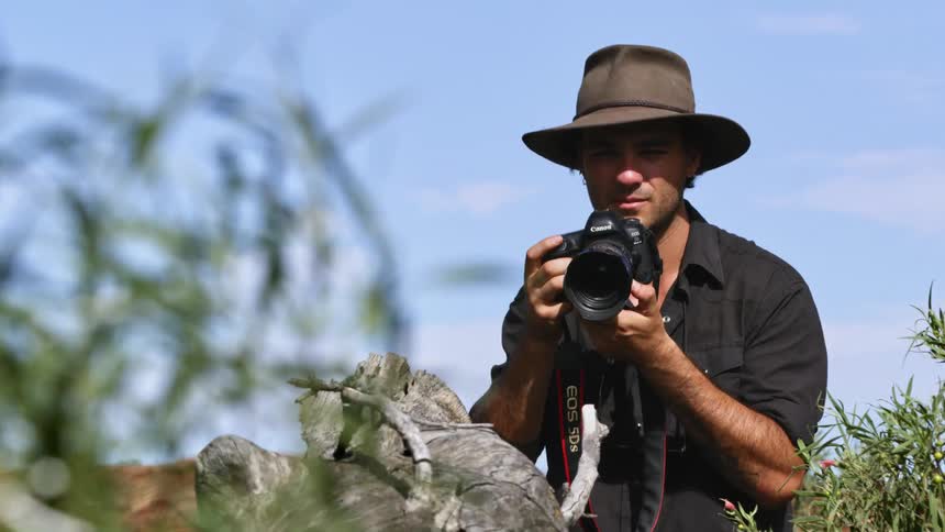A man with a hat and a camera taking pictures with the sky and clouds in the background