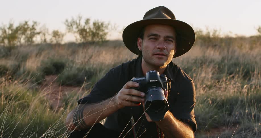 A man with a hat and camera taking pictures with grass and the sky in the background