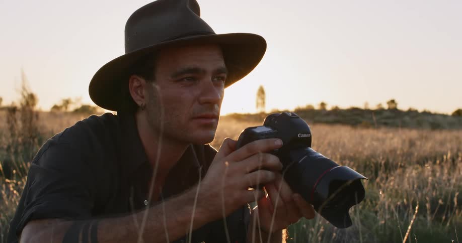 A photographer kneels in the tall grasses of a grassland and raises his camera to take a photograph