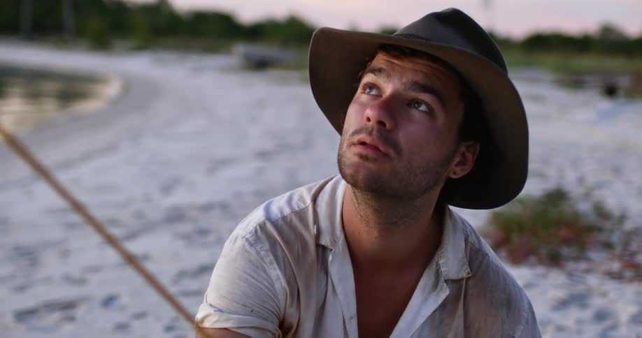Man wearing hat ties a rope to a stake on the sand of a beach at sunset