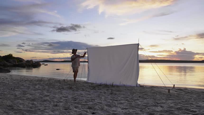 A man smooths out a sheet being used as a makeshift tent on the beach next to the water