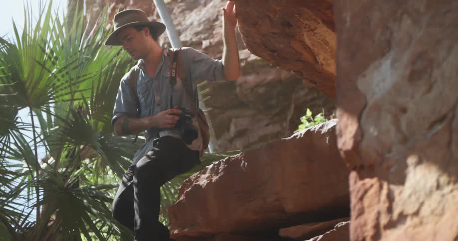A young man carrying a camera climbs down a cliff