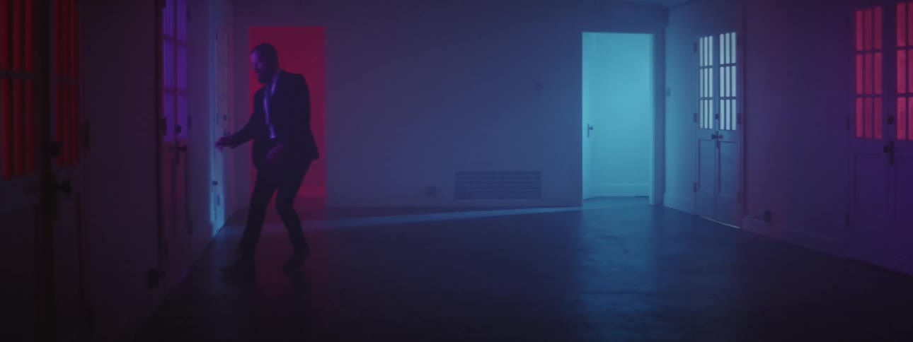 A man runs from a blue illuminated doorway through a room illuminated by purple light to a door which he quickly opens and runs through