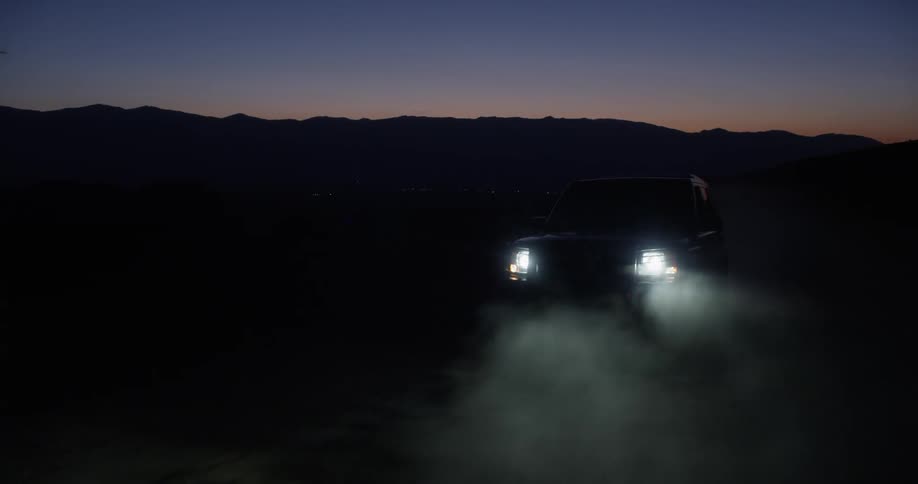 A car drives down a dusty road with headlights on at dusk