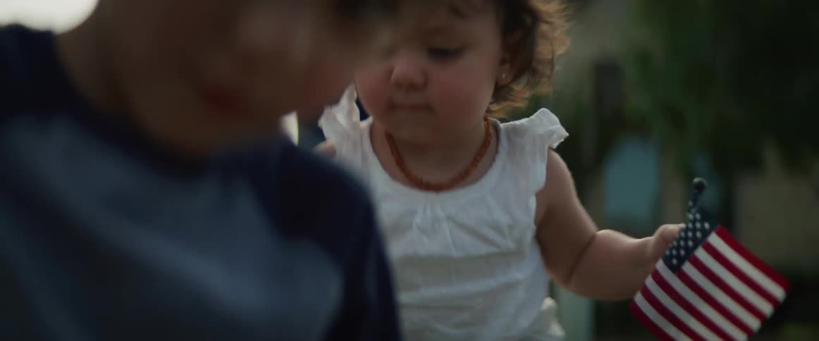 Little girl holds two american flags as she sits outdoors next to a little boy while their mother watches