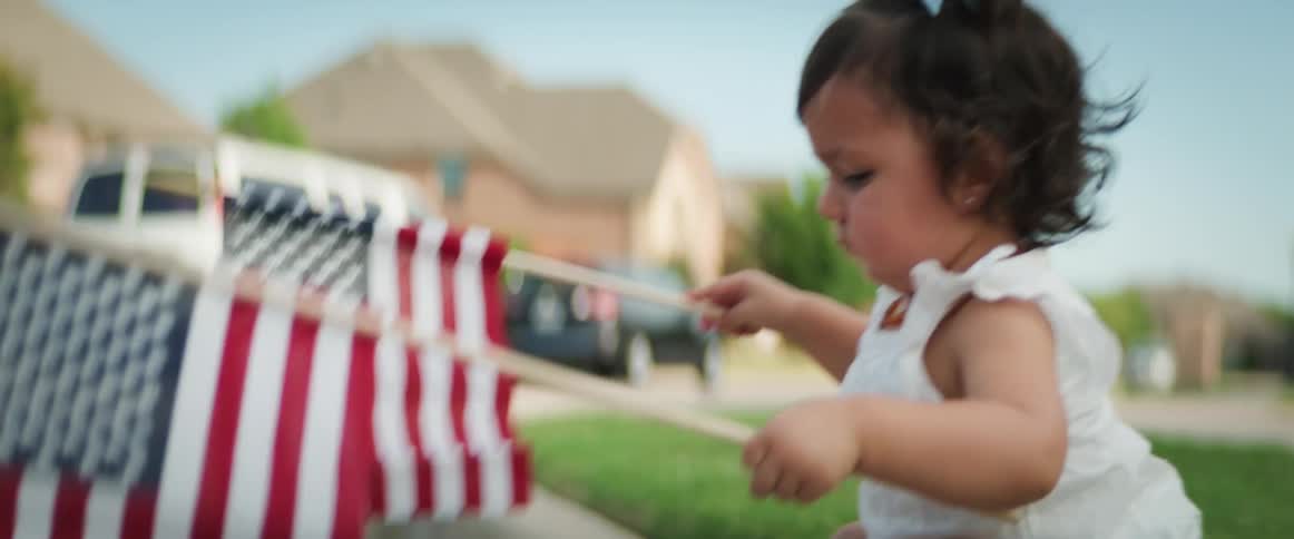 A little boy walks past his baby sister who hold two small american flags outside