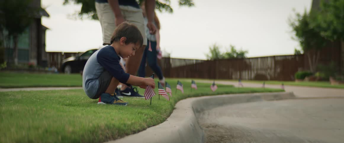 A man hands a small flag to his son and the two plant the flags in the grass next to a curb as a woman and little girl follow behind