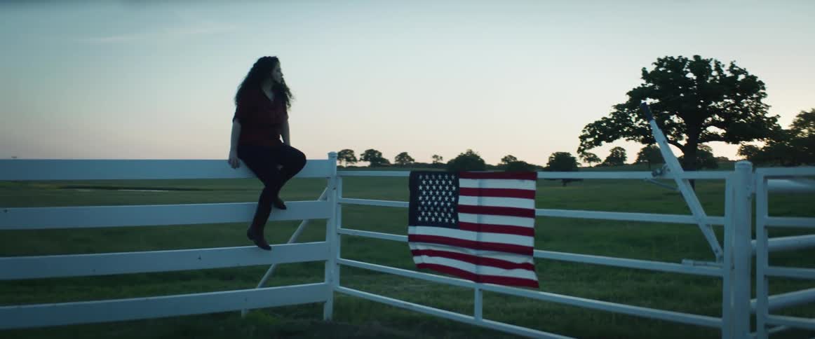 A woman sits on a fence next to a gate with an american flag hanging from it in a vast field of grass