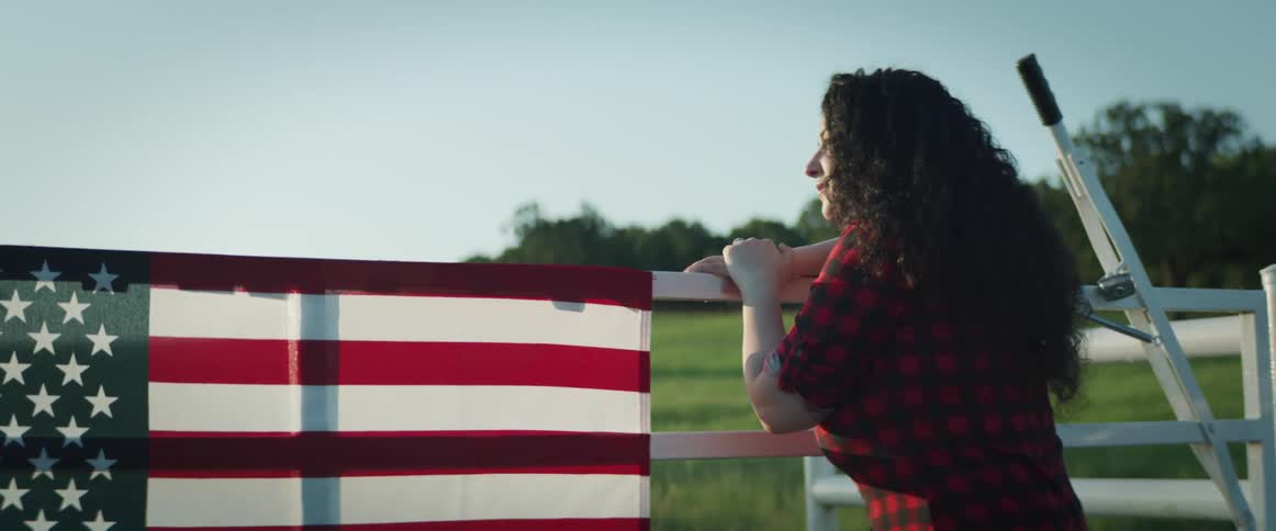 Woman walks up to a fence and leans against it as an american flag hangs next to her