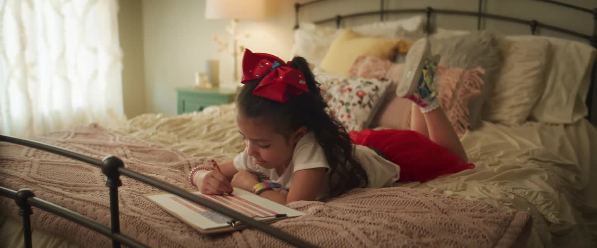A young girl lies on a bed coloring a picture of an american flag on a clipboard