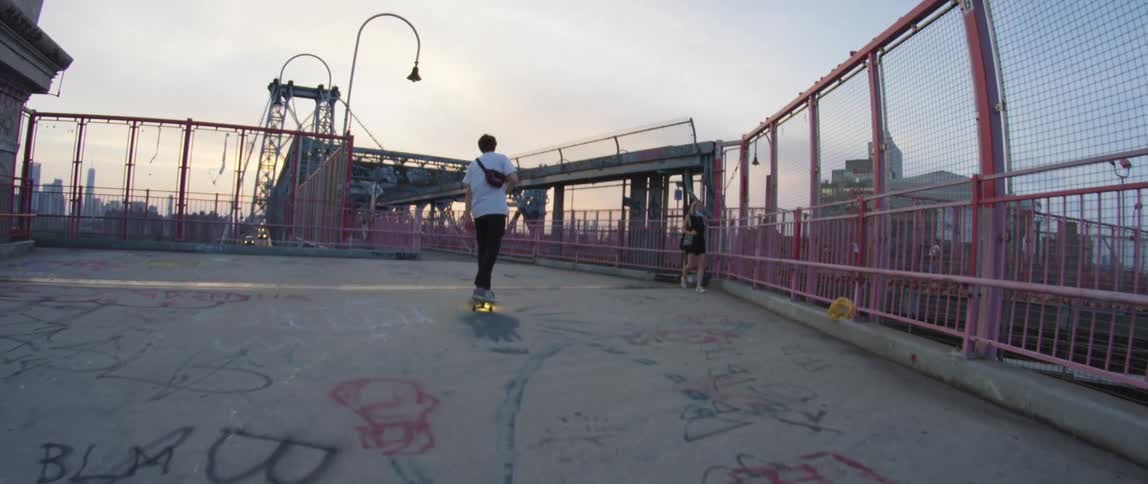 A teenage boy skateboards past a girl on a bridge with graffiti