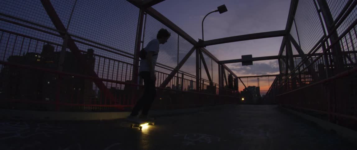 A teenage boy skateboards across a bridge with a city skyline in the distance