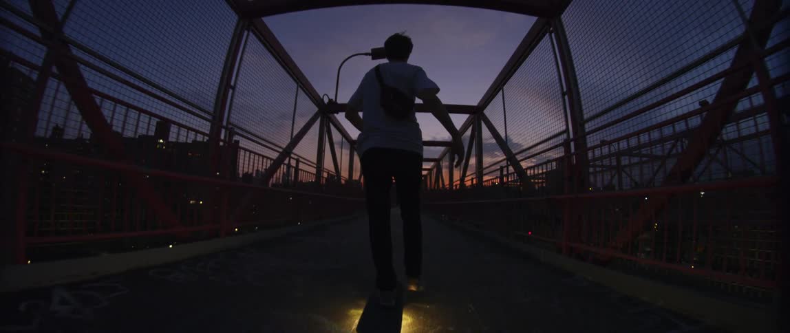A teenager skateboards across a bridge with a city skyline in the background at dusk