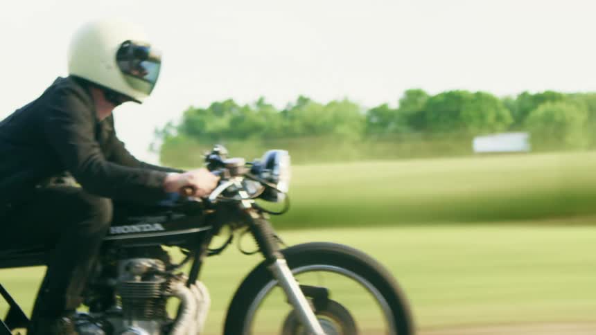 A man driving his motorcycle down a street past a grassland