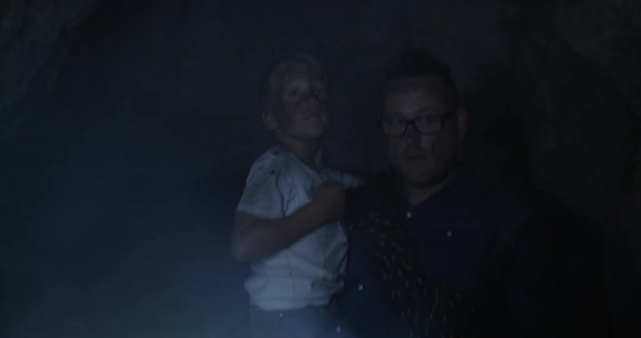 A man carrying a flashlight and holding a young boy as he is walking through a cave