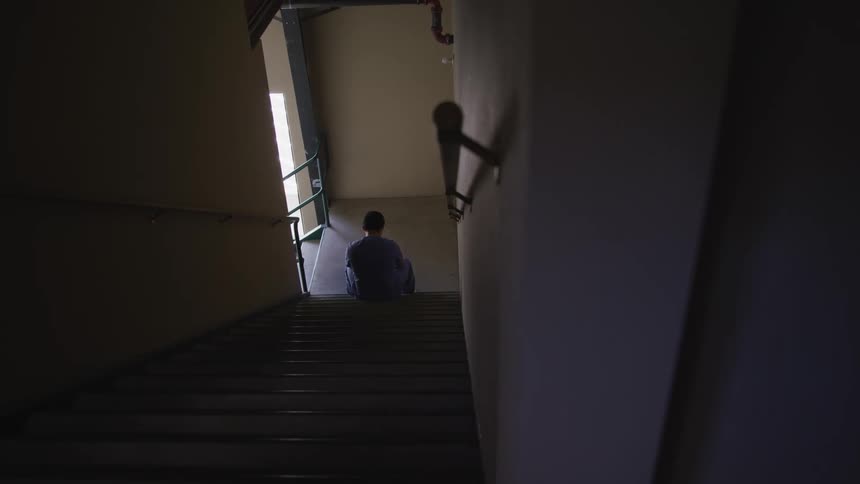 A tired nurse wearing blue scrubs sits on the dimly lit stairs of a hospital