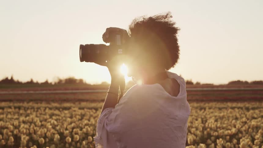 Black woman with curly hair and a white shawls smiles broadly and takes photographs with a camera at sunset in tulip fields