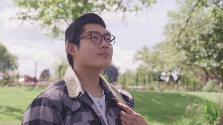 A young asian man stands outdoors on a sunny day and looks up at the sky while carrying a backpack