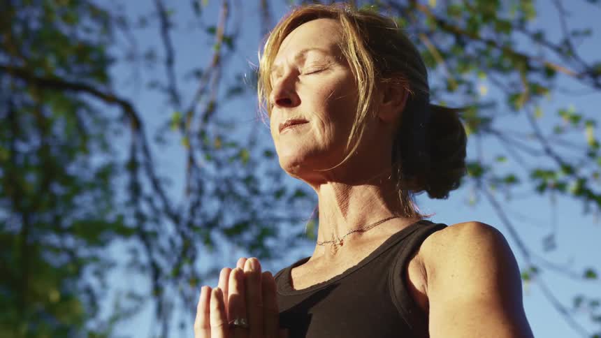 Middle aged blonde woman puts her hands together under her chin and stands with eyes closed under tree branches