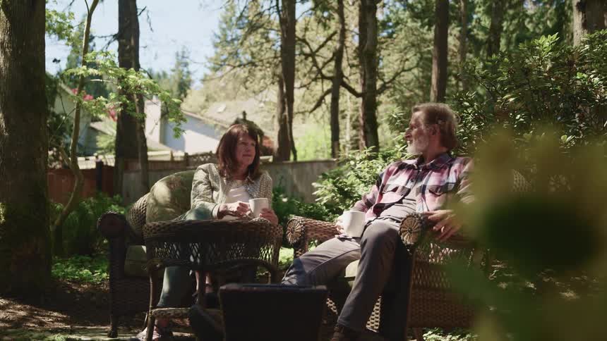 Middle aged man with a beard and woman with brown hair sitting in armchairs drink two cups of coffee in the garden