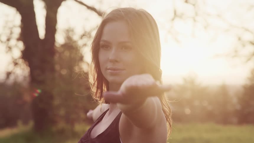 A woman holds a yoga pose as she stands with her arms raised in a park