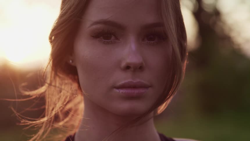 Woman with brown hair and makeup faces the camera with eye contact at sunset