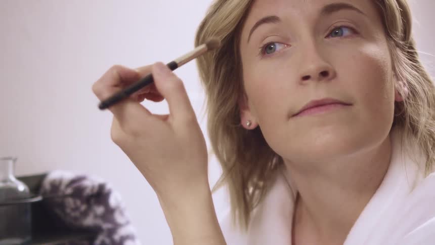 Woman wearing white robe applies makeup on her face using a brush