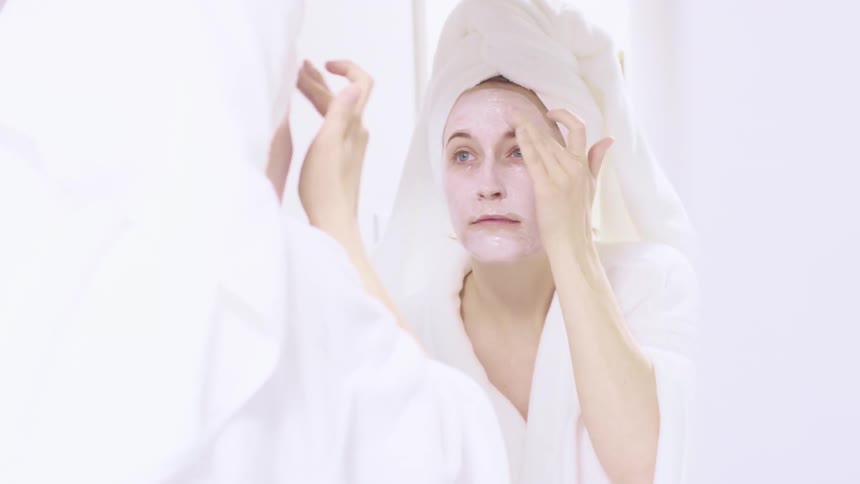 A woman stands in front of her mirror and puts cream on her face