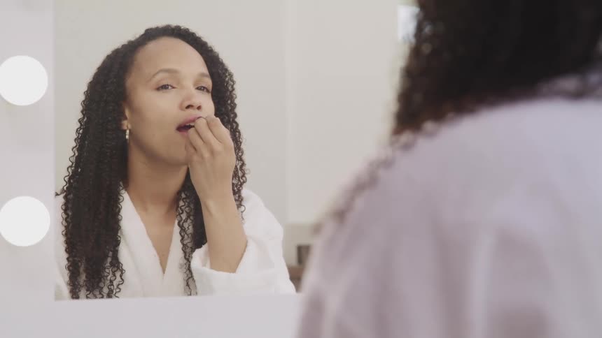 Woman with curly hair and a white bathrobe looks in the mirror and applies lipstick in bathroom
