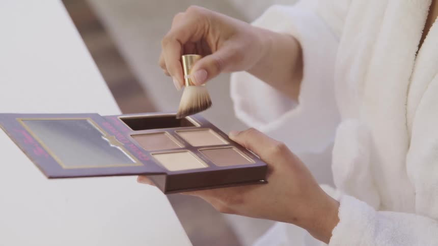 Black woman holds a makeup tray and applies makeup with a brush as she sits on the edge of a bed
