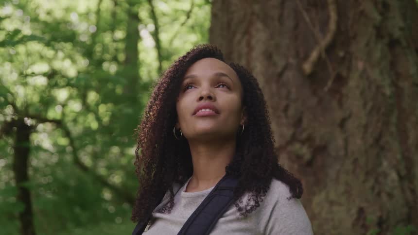 A backpacker looks up at the sky in the forest and smiles as she closes her eyes